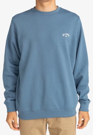 Sudadera de forro polar azul con cuello redondo, puños y dobladillo acanalados, que presenta un pequeño logo blanco en el lado izquierdo del pecho. Textura suave.