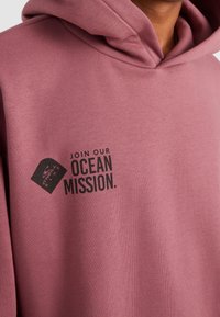 Sudadera con capucha en un tono rosa apagado, con un logotipo gráfico y el texto "ÚNETE A NUESTRA MISIÓN OCEÁNICA" impreso en negro. Textura de material suave.