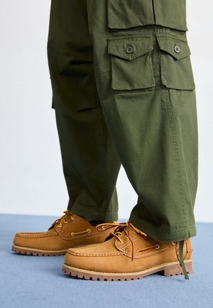 Helle Lederschuhe mit gelben und kastanienbraunen Schnürsenkeln. Olivgrüne Cargo-Hosen mit Seitentaschen, lockerer Passform und an den Knöcheln gebundenen Bündchen.