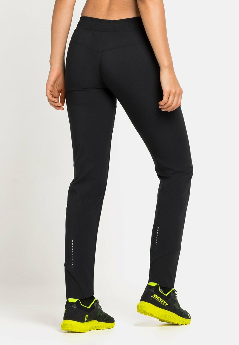 Schwarze Sporthosen mit glattem Material, tapered Fit, elastischem Bund und reflektierenden Details an den unteren Beinen. Kombiniert mit schwarzen und gelben Schuhen.