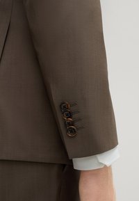 Ärmel eines braunen Sakkos mit fünf dunklen Knöpfen und braunen Akzenten, glatte Stoffoberfläche und ein sichtbares weißes Hemd. 