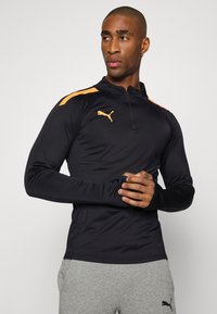 Schwarzes, langärmeliges Sportoberteil mit orangen Akzenten und einem Viertel-Reißverschluss-Kragen; verfügt über ein Puma-Logo auf der Brust. Getragen mit grauen Sweatpants.