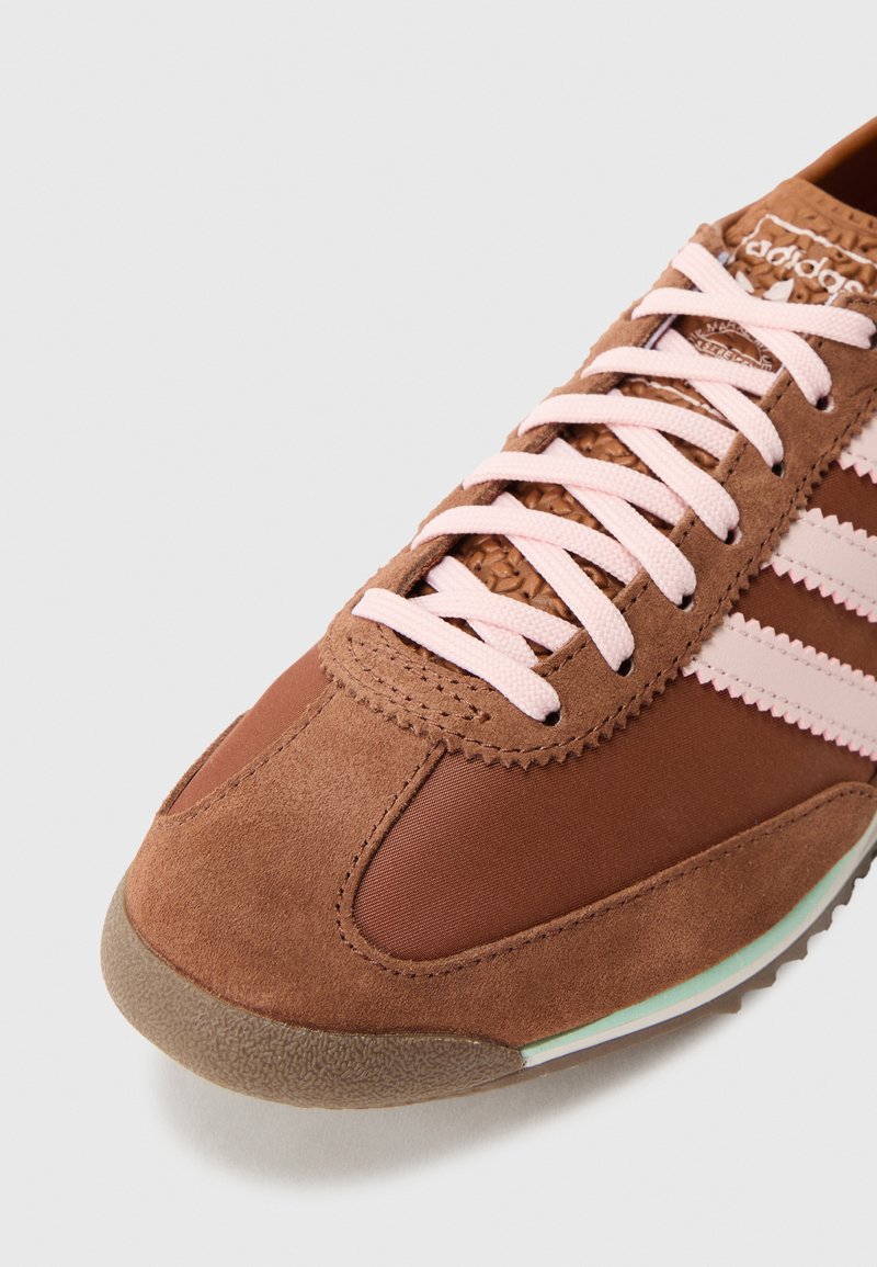 Brown suede and fabric sneaker with pink laces and stripes, textured toe cap and part of the sole visible on a white background.