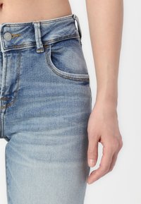 Jean en denim bleu clair taille haute avec une finition délavée, featuring une poche avant unique et des détails de couture visibles.