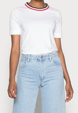 Nainen, joka pukeutunut valkoiseen lyhythihaiseen paitaan, jossa on punaisen ja mustan raidallinen kaulus, sekä vaaleansiniset korkeavyötäröiset farkut.