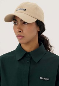 Beżowa czapka z zakrzywionym daszkiem ma czarne logo "MISAGA®". Model nosi ciemnozieloną koszulę z kołnierzykiem, zapinaną na guziki i o subtelnej teksturze.
