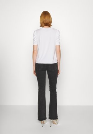 Person mit kurzen roten Haaren, die ein schlichtes weißes kurzärmeliges Shirt, schwarze Jeans und beige High Heels trägt und mit dem Rücken zur Kamera steht.