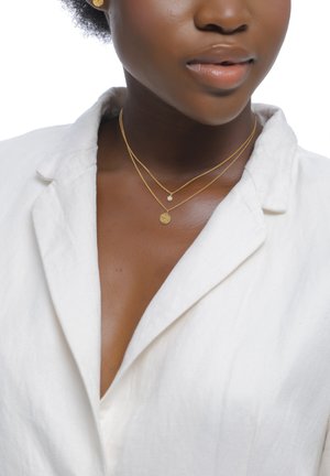 Gros plan d'une personne portant une veste blanche et deux colliers en or délicats : l'un avec un petit pendentif en diamant, l'autre avec un charme rond texturé.