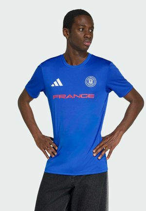 Mann mit Händen in der Hüfte, der ein blaues französisches Sporttrikot mit Adidas-Logo und Emblem auf der Brust trägt und zur Seite schaut.