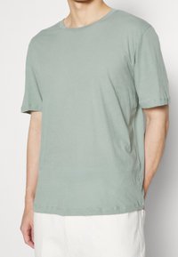 T-shirt en coton vert clair avec un col rond, des manches courtes, une coupe décontractée et des détails de couture visibles le long de l'ourlet et des manches.