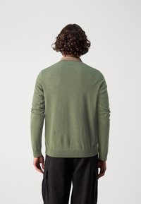 Zelený pletený kardigan s dlouhými rukávy, vroubkovaným lemem a kontrastním hnědým límcem. Hladká textura, přiléhavý střih, nošený přes černé cargo kalhoty.