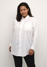 Camisa blanca de botones hecha de tela suave, con un collar clásico, mangas largas y un bolsillo frontal. Usada con pantalones negros.