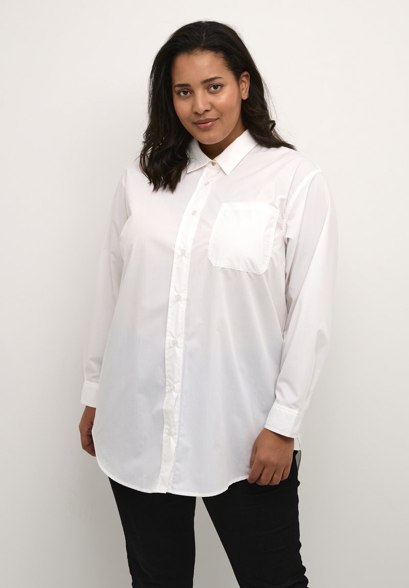 Camisa blanca de botones hecha de tela suave, con un collar clásico, mangas largas y un bolsillo frontal. Usada con pantalones negros.