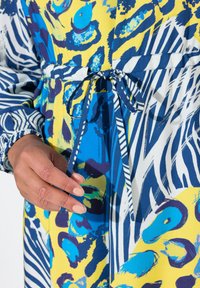 Kleurvolle katoenen jurk met gedurfde blauwe en gele abstracte patronen, met een strik in de taille en marineblauwe- en witte gestreepte accenten.