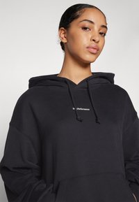 Svart hoodie med en framficka och snörning i huven. Har vit logotext på bröstet, tillverkad av mjukt tyg med en avslappnad passform.