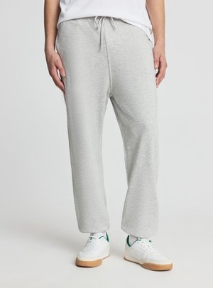 Osoba ubrana w jasnoszare spodnie dresowe ze sznurkiem i białe sneakersy z zielonymi akcentami, stojąca na jednolitym tle.