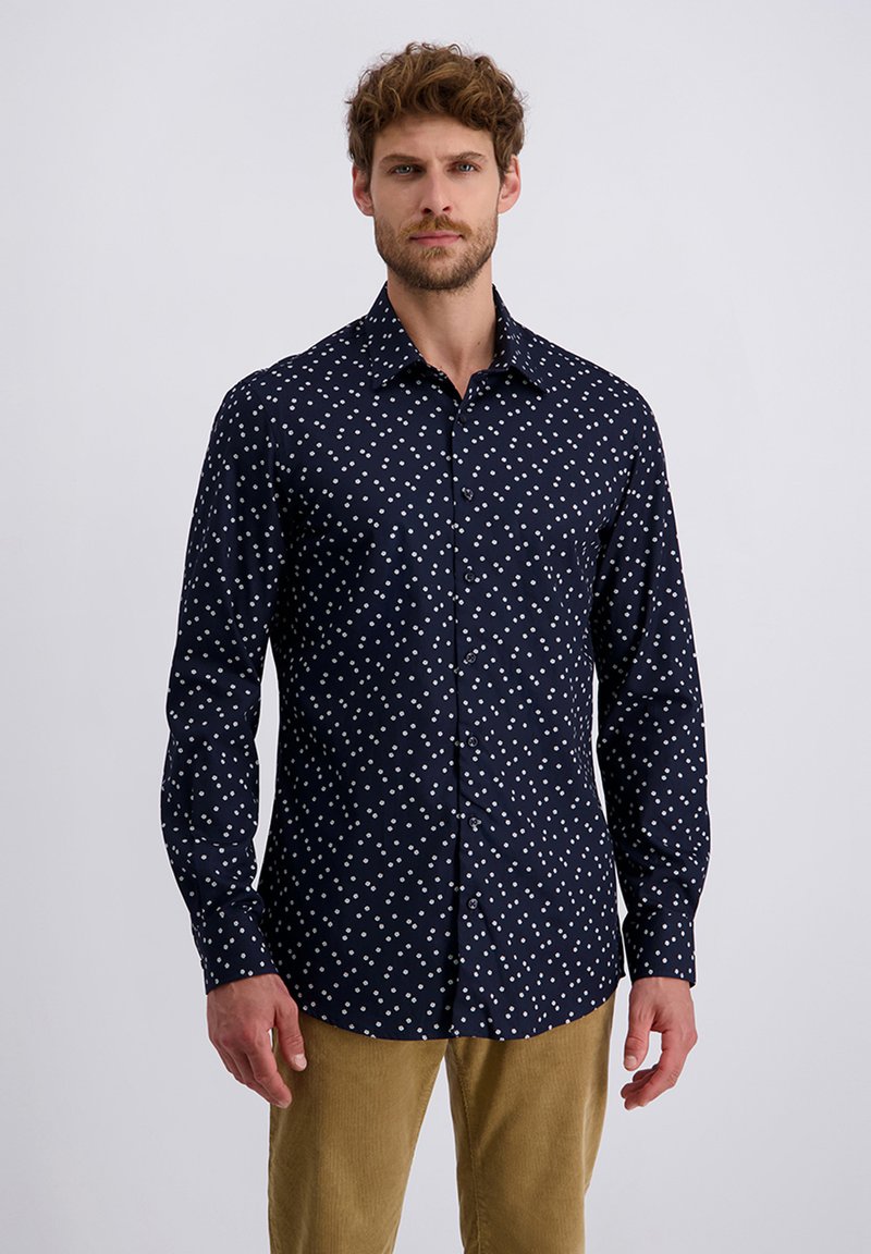 Mann in einem marineblauen Langarmhemd mit weißen Punkten und hellbraunen Hosen, der vor einem schlichten hellgrauen Hintergrund steht.