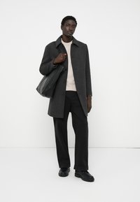 Férfi sötét kockás kabátban, világos pulóverben, fekete nadrágban, fekete cipőben, fekete bőr táskával a kezében, fehér háttér előtt.