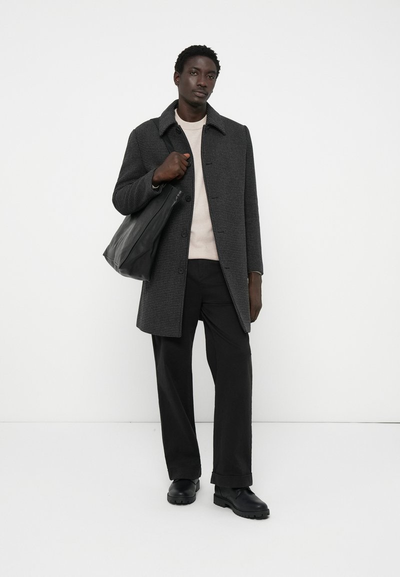 Homme portant un manteau à carreaux sombres, un pull léger, un pantalon noir, des chaussures noires, tenant un sac fourre-tout en cuir noir sur fond blanc.