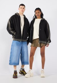 Felpe nere con zip e effetto sfumato, abbinate a una maglietta beige. Un modello indossa jeans blu larghi, l'altro una gonna a quadri con sneakers.