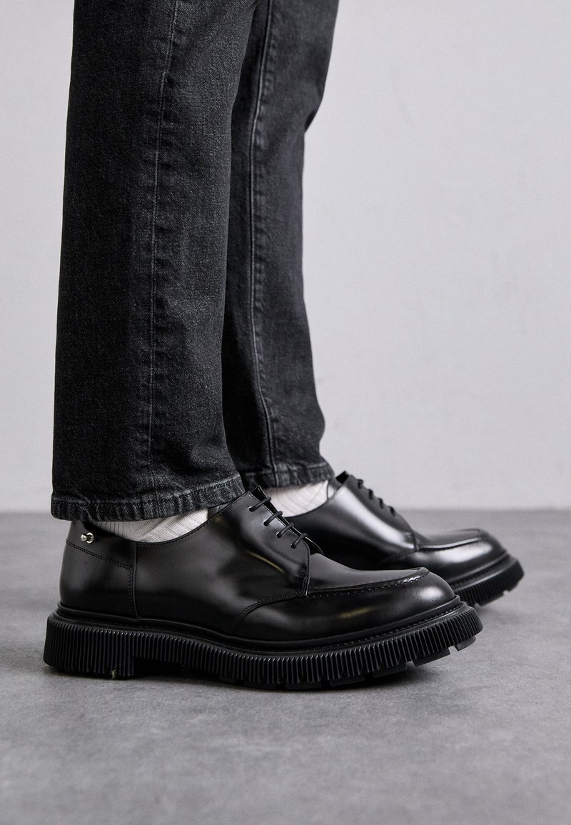 Mustat nahkakengät, joissa on sileä pinta ja paksu, uritettu kumipohja. Nauhat ja hienovarainen metallikoriste ovat näkyvissä. Yhdistetty tummiin farkkuihin.