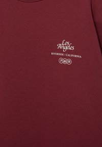Bordeauxrode stof met witte tekst met de woorden "Los Angeles," "Riverside / California," en het "P&B"-logo in het midden van het borstgebied.