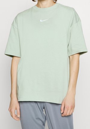 Person in locker grünem Nike-T-Shirt mit weißem Logo und grauer Hose, vor einem einfarbigen hellen Hintergrund stehend.