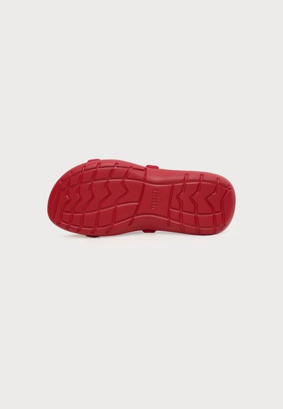 Semelle de chaussure en caoutchouc rouge vue par dessous, montrant des crampons texturés en motifs zigzag et grille, centrée sur un fond clair uni.