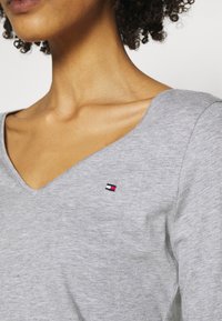 Grijze v-hals top van zachte stof, met een klein geborduurd logo in rood, wit en blauw op de linkerborst.