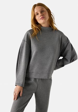 Sudadera gris recortada con cuello alto y mangas anchas, hecha de una tela suave. Combinada con pantalones grises a juego. Detalles de costura minimalistas.