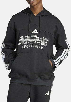 Persona che indossa una felpa sportiva nera Adidas con logo bianco e strisce sulle maniche, mani nella tasca frontale, sorriso leggero.