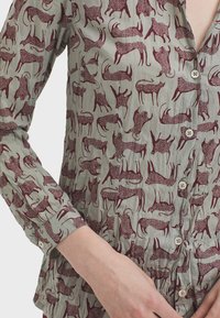 Camicia a maniche lunghe in tessuto verde chiaro con un motivo animale color marrone. Chiusura con bottoni e piccoli bottoni rotondi, con dettagli arricciati sull'orlo.