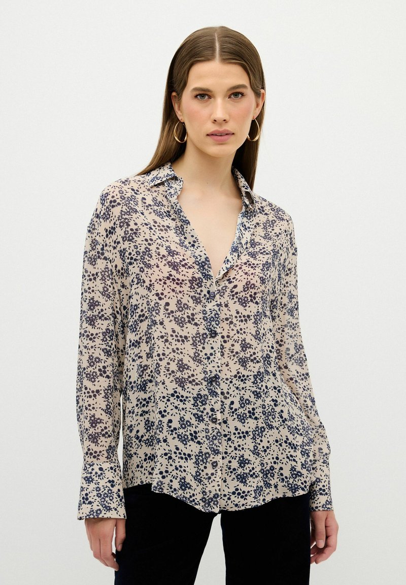 Blusa ligera y transparente en beige con patrones florales en azul marino. Cuenta con un collar clásico, cierre de botones en la parte delantera y mangas largas con detalle en los puños.