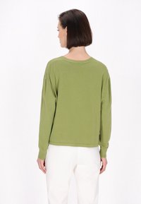 Zöld, hosszú ujjú pulóver laza fazonban, bordás mandzsettákkal és aljjal. Fehér nadrággal viselve, a puha, texturált anyag hangsúlyozásával.