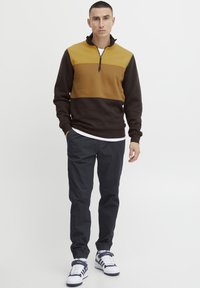 Fleece-Pullover mit einem Viertelreißverschluss-Kragen, der ein braunes und senfgelbes Farbblockdesign aufweist. Kombiniert mit dunklen Hosen und weißen Sneakers.