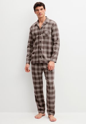 Man staat blootsvoets en draagt een bruin-beige geruit overhemd met lange mouwen en knopen en bijpassende pyjamabroek tegen een witte achtergrond.