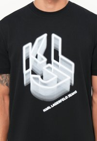 Melnas kokvilnas T-krekls ar baltu, 3D grafiku, kurā attēloti burti "KL" un "KARL LAGERFELD JEANS", drukāti mūsdienīgā tipogrāfijas stilā.