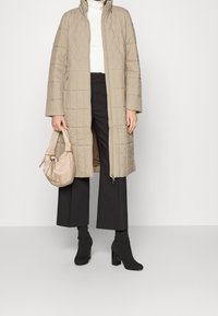 Abrigo largo acolchado beige con cuello alto, cierre de cremallera y dos bolsillos laterales. Combinado con pantalones negros de pierna ancha y botines negros puntiagudos.