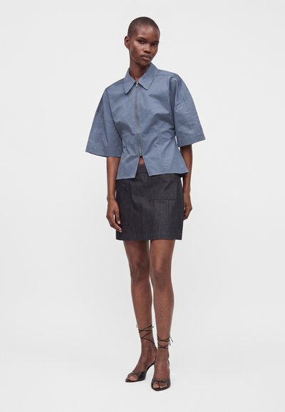 Femme portant une chemise bleue à manches courtes zippée, une jupe noire au-dessus du genou et des sandales noires à talons hauts à brides, debout sur un fond blanc.
