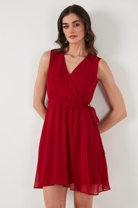 Woman wearing sleeveless red wrap dress with waist tie, standing against a white wall, looking slightly to the side.