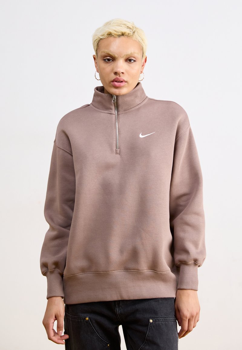 Felpa Nike con cappuccio di colore marrone chiaro, con colletto alto, mezza zip, maniche lunghe e polsini e orlo a coste. Presenta il logo bianco sul petto.