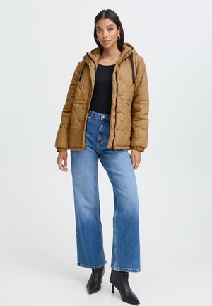 Femme debout portant une veste matelassée marron à capuche, un haut noir, un jean bleu à jambes larges et des bottes noires à talons pointus devant un fond uni.