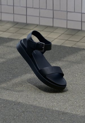 Sandale en cuir noir avec bride cheville et boucle, semelle coussinée, présentée devant un mur carrelé et un sol en asphalte.