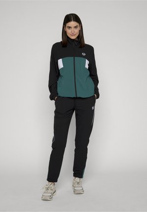 Kvinne står med hendene i lommene, iført svart og grønn jakke med glidelås, svarte bukser med hvite striper og lyse joggesko.