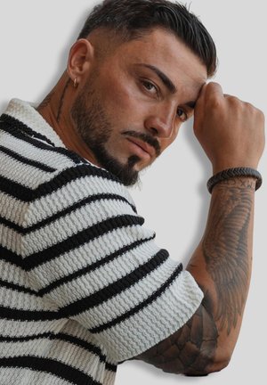 Man with short dark hair and beard wearing a black and white striped shirt, resting his arm with a wing tattoo and braided bracelet near his face.