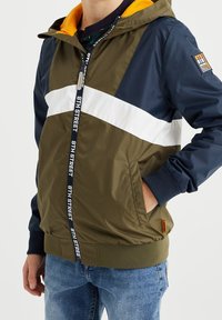 Vítr bránící bunda v olivově zelené, námořnické modré a bílé barvě s kapucí, zipovým zapínáním a elastickými manžetami. Obsahuje dekorativní štítek na zipu.