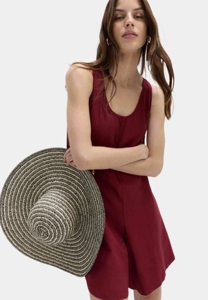 Mujer vistiendo un vestido sin mangas color burdeos, sosteniendo un gran sombrero de paja blanco y negro, con los brazos cruzados frente a un fondo liso.