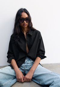 Camicetta nera con maniche arrotolate, abbinata a jeans chiari strappati. Occhiali da sole tartarugati di grandi dimensioni completano il look.