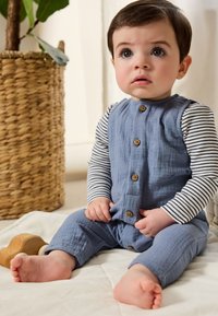 Ensemble en coton bleu avec un devant boutonné texturé. Chemise à manches longues rayée sous le haut sans manches. Enfant assis, pieds nus.