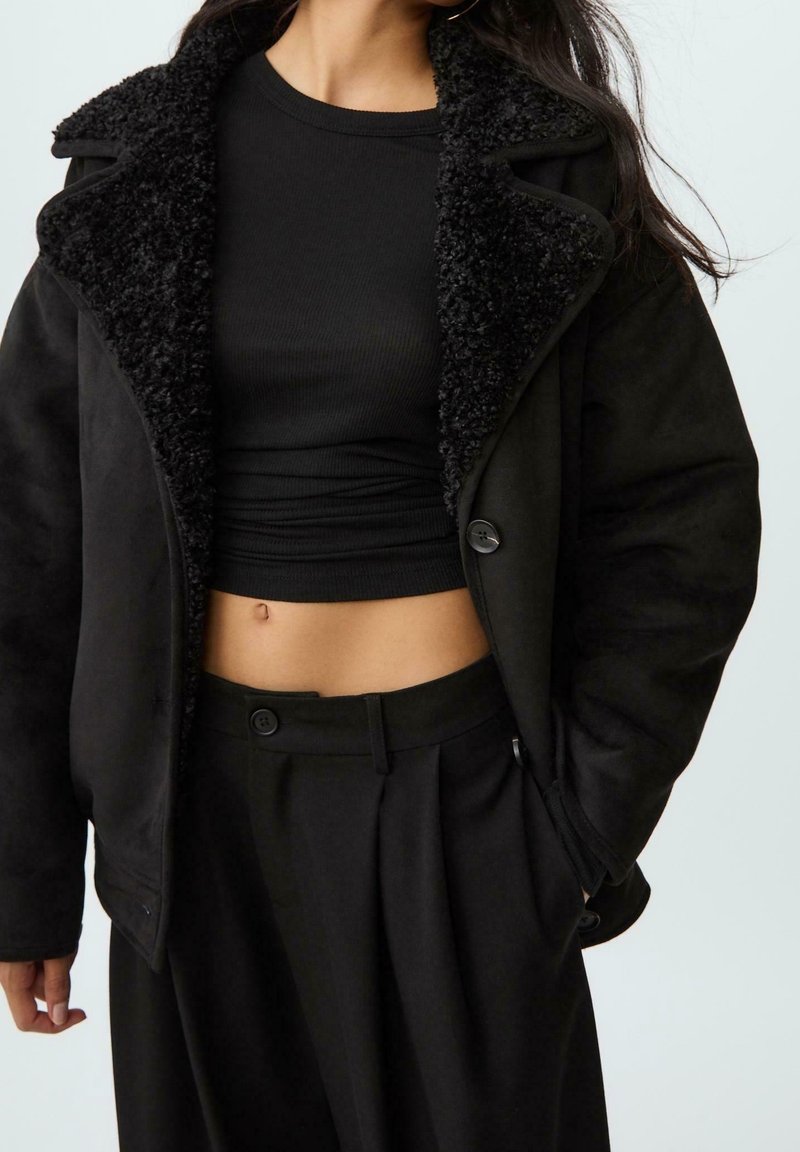 Femme portant un top court noir côtelé, un pantalon noir taille haute et une veste noire doublée en peau de mouton, une main dans la poche.